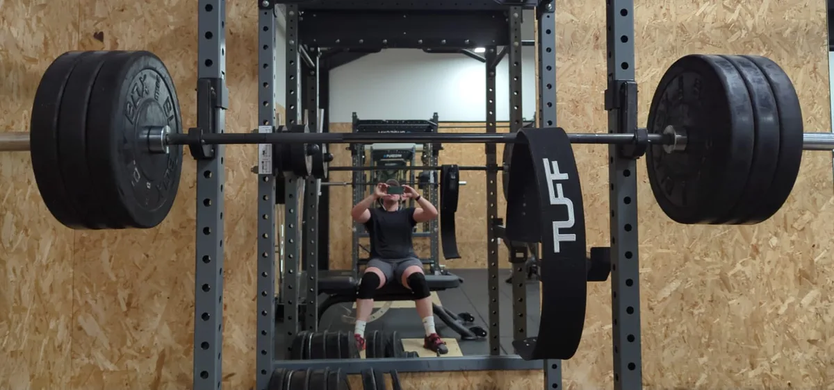 Graham in the gym, seen in the mirror behind a loaded barbell